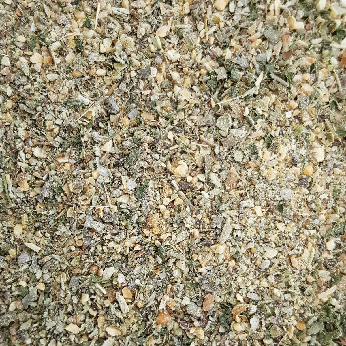 A close-up, top-down view of a coarse, granular herb and spice blend containing dried green leaves, beige seeds, and light brown roots mixed together.