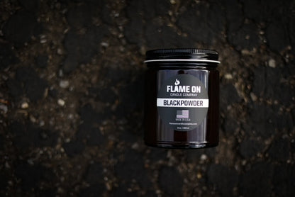 A dark colored candle with a label reading 'Flame On Black Powder', placed on an asphalt surface.