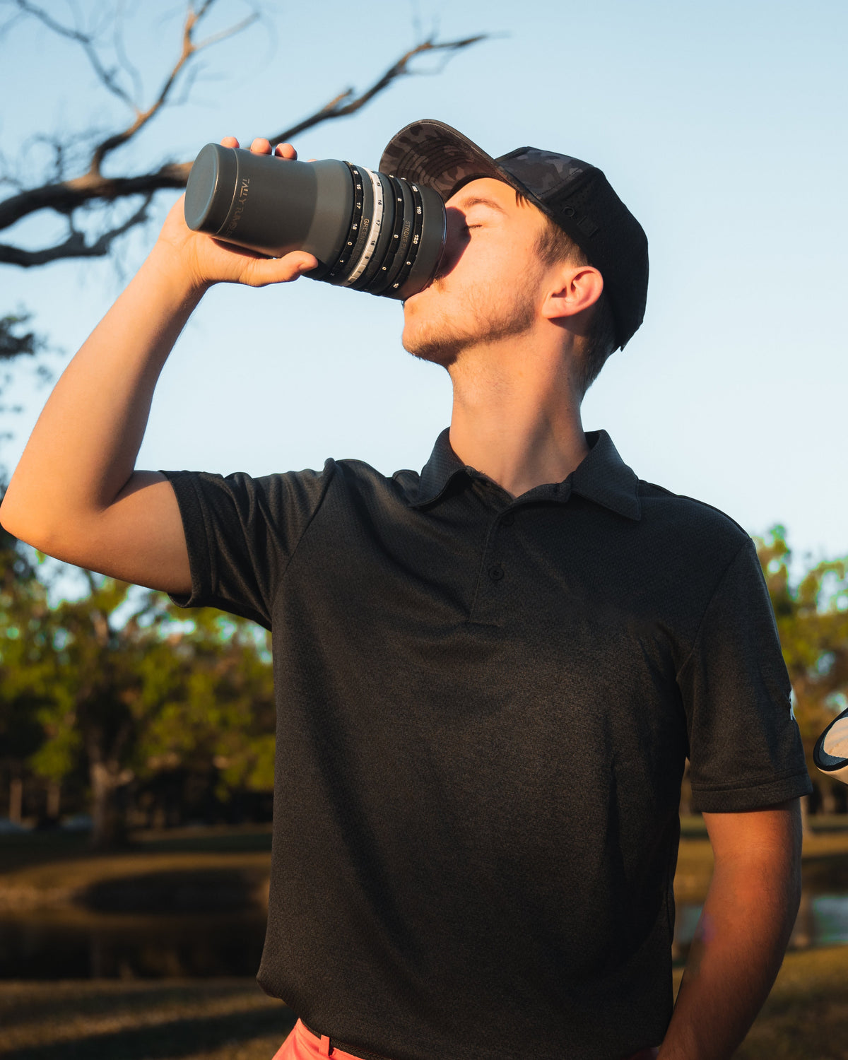 Man drinking a tally tumbler outdoors with trees and sky in the background
