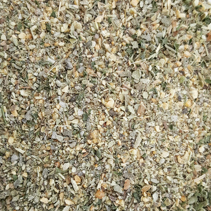 A close-up, top-down view of a coarse, granular herb and spice blend containing dried green leaves, beige seeds, and light brown roots mixed together.
