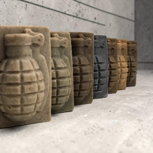 A row of rectangular soap bars shaped like hand grenades, shown in muted earthy tones including beige, brown, gray, and tan, arranged diagonally on a light concrete surface against a textured gray wall.
