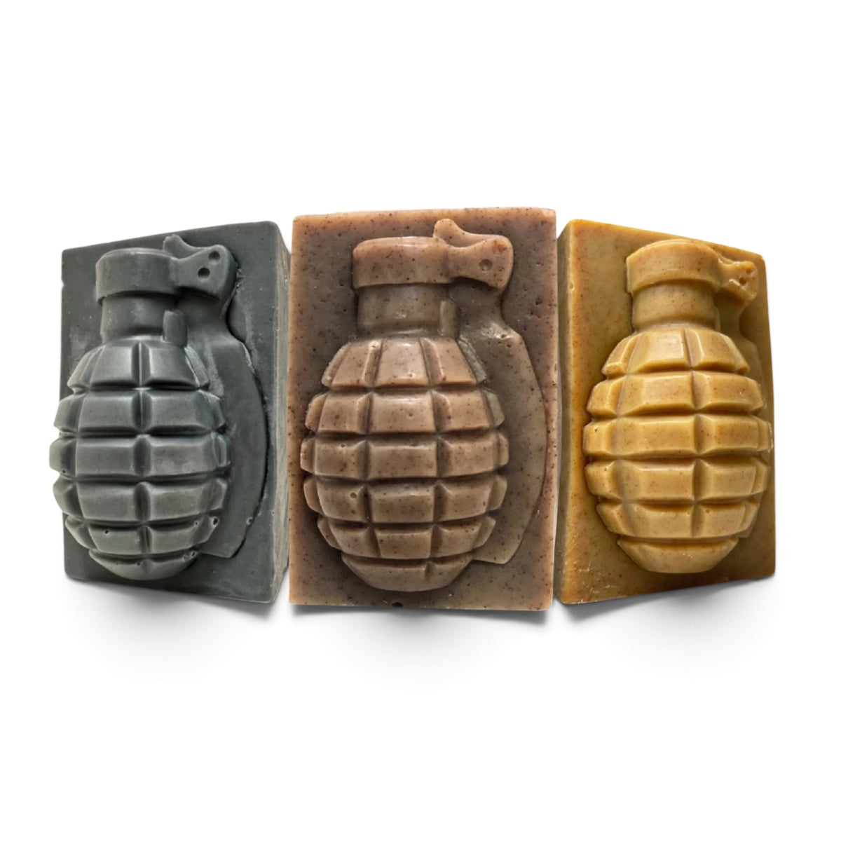 Three rectangular soap bars shaped like hand grenades, shown side by side on a white background. The left soap is dark gray, the middle is a warm taupe-brown, and the right is a mustard yellow.