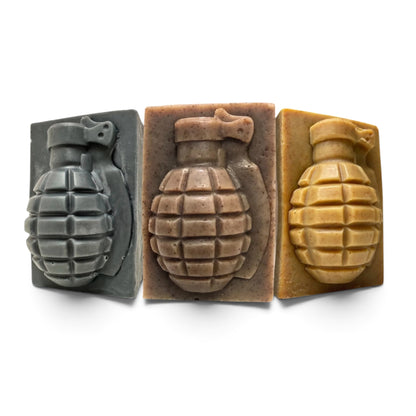Three rectangular soap bars shaped like hand grenades, shown side by side on a white background. The left soap is dark gray, the middle is a warm taupe-brown, and the right is a mustard yellow.