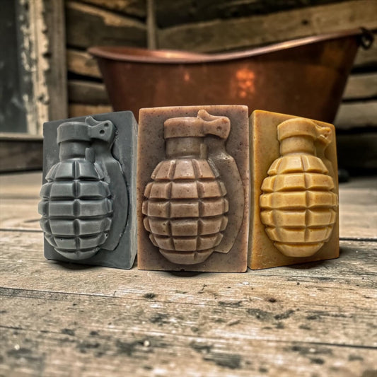 Three rectangular soap bars shaped like hand grenades are displayed on a rustic wooden surface. The soap bars come in three different colors: dark gray, medium brown, and tan.