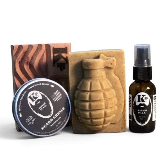 A beard grooming set on a white background, including a tin of K&H Beard Grease, a hand-crafted soap bar molded like a grenade, a small amber spray bottle of K&H Hair Care, and a wooden box.