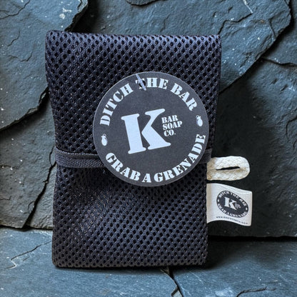 A black mesh soap bag with a black elastic band and a round black label reading 'Ditch The Bar Grab A Grenade' and 'K Bar Soap Co.' tied with a white string, set against a dark slate background.