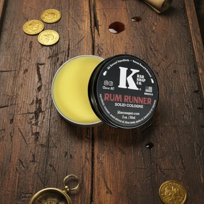 Solid cologne tin labeled 'Rum Runner' by K Bar & Brush Co on a wooden surface with gold coins.