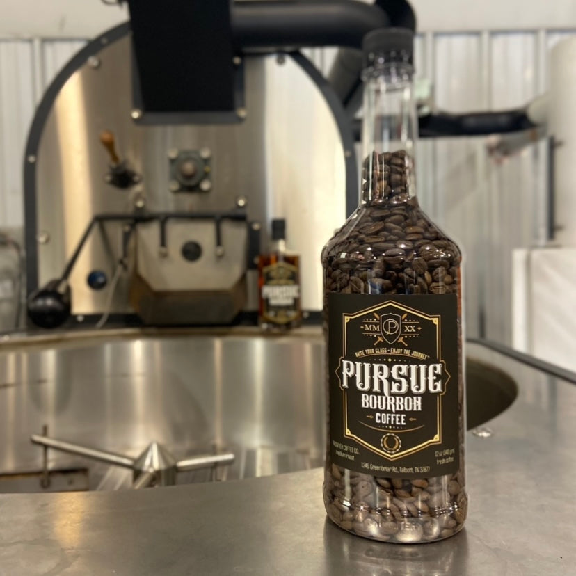 A bottle of Pursue Bourbon Coffee on a counter with a coffee roasting machine in the background.