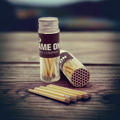 A match jar with the label 'Flame On Candle Company' next to a scattered row of matches on a wooden surface.