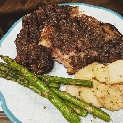 A close-up of a cooked steak with a dark, seasoned crust, served on a light blue patterned plate with green asparagus and sliced potatoes.