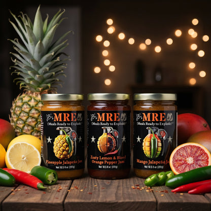 Three jars of MRE brand hot chili peppers arranged on a wooden table with fresh fruit and peppers around them, including pineapple, mango, citrus, jalapeños, and red chiles, against a dark background with warm string lights.