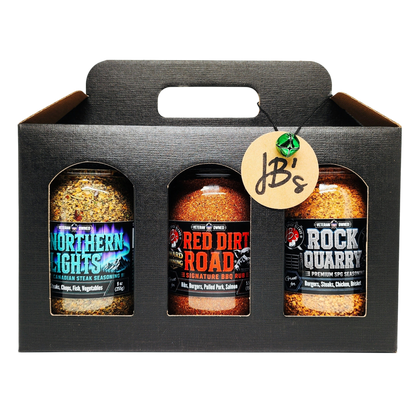 A three-pack of JB's smoked meats seasoning bottles in a black cardboard carrier. The bottles are labeled Northern Lights, Red Dirt Road, and Rock Quarry, and are shown against a clean white background.