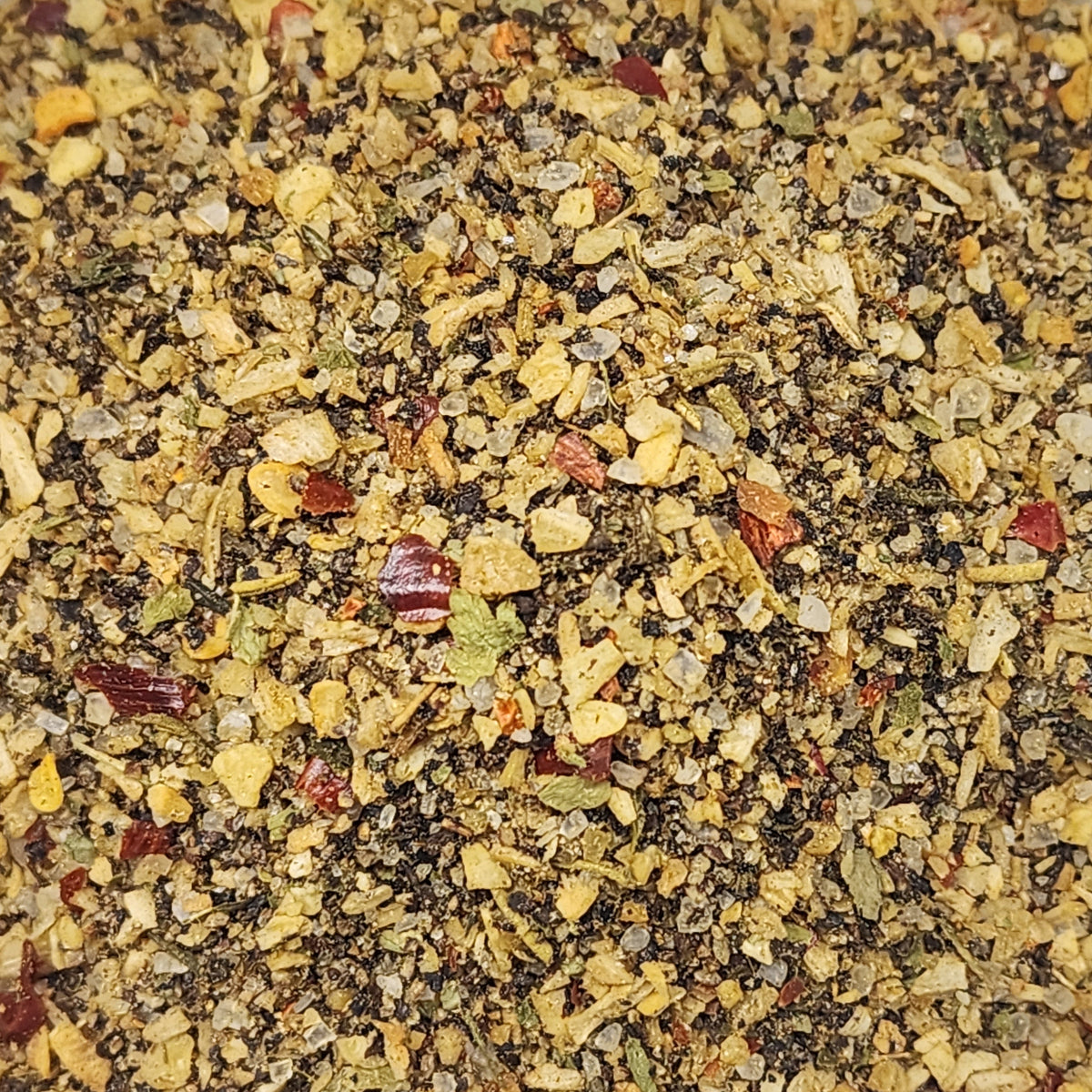 A close-up, top-down view of a textured spice blend containing ground black pepper, dried herbs, garlic, and red chili flakes.