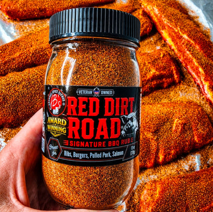 A hand holds a jar of Red Dirt Road Signature BBQ Rubie seasoning in front of a close-up background of food coated with the orange-brown spice rub.